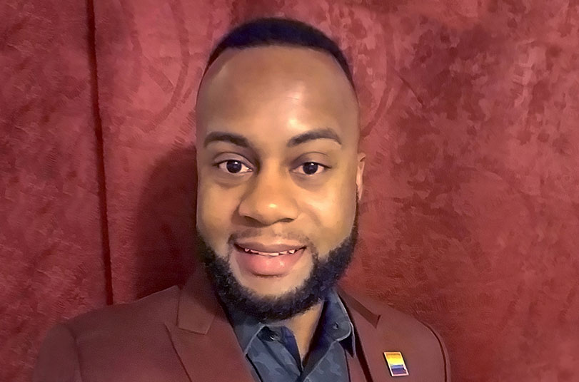 Luzerne smiling in a maroon suit with a rainbow Pride pin. | LGBTQ
