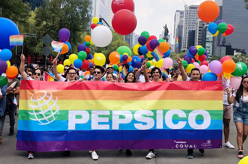 Mario marches at a Pride parade with friends and coworkers holding a giant Pepsico Pride banner as rainbow balloons and flags bounce behind them. | LGBTQ