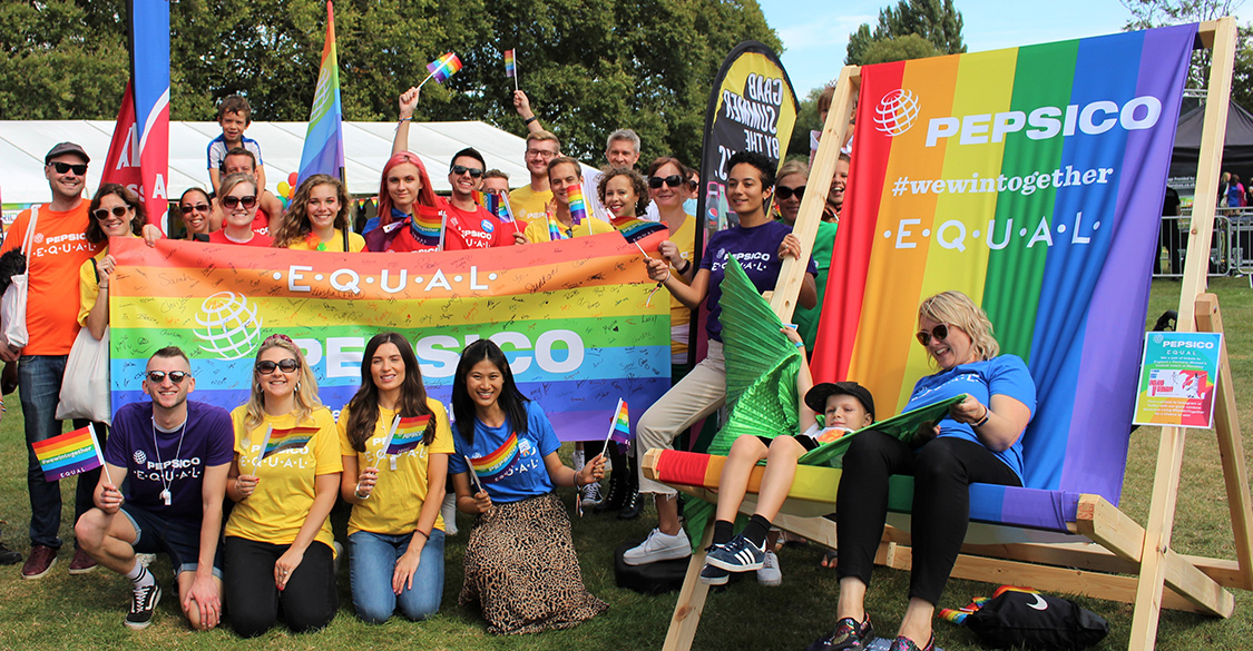 Lively group of 25 men, women, and children outdoors and in rainbow attire celebrating Pride at Pepsi with banners and flags.