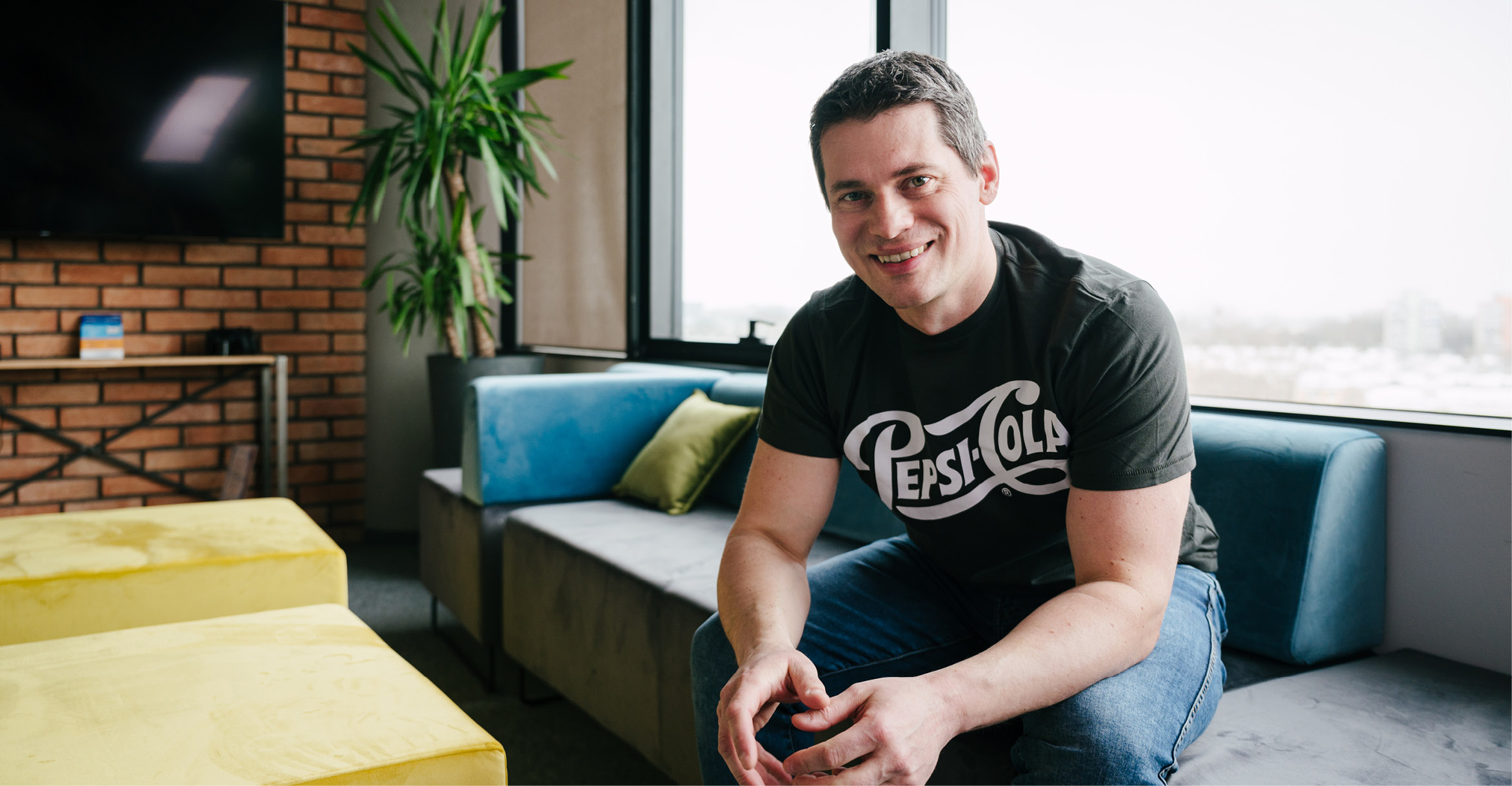 Filip Sielka sits on a couch inside his brightly lit workspace with colorful blue, green and yellow furnishings. He has short brown hair and is wearing jeans with a PepsiCo t-shirt. He smiles as he casually leans forward resting his elbows on his knees. | GBS