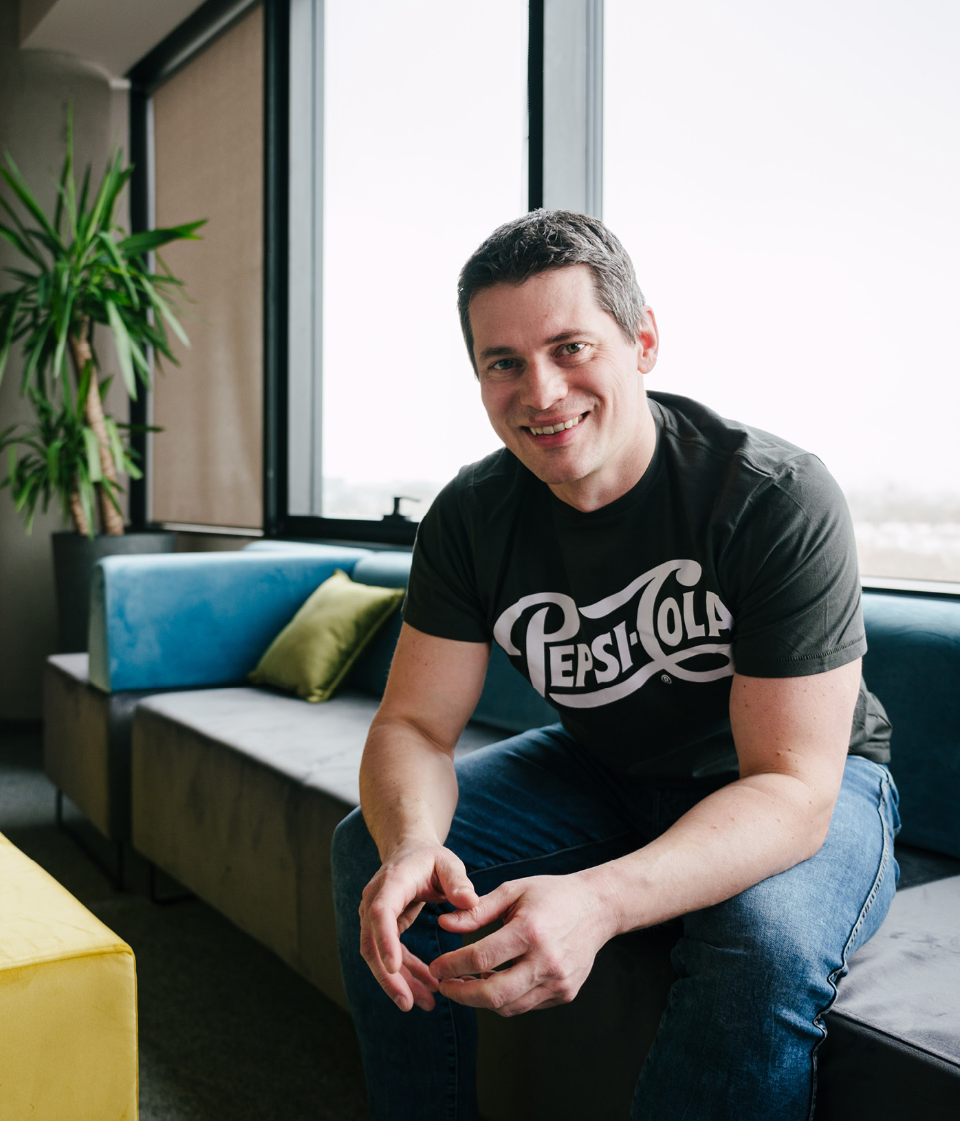 Filip Sielka, GBS team lead, sits on a couch inside his brightly lit workspace with colorful blue, green and yellow furnishings. He has short brown hair and is wearing jeans with a PepsiCo t-shirt. He smiles as he casually leans forward resting his elbows on his knees.